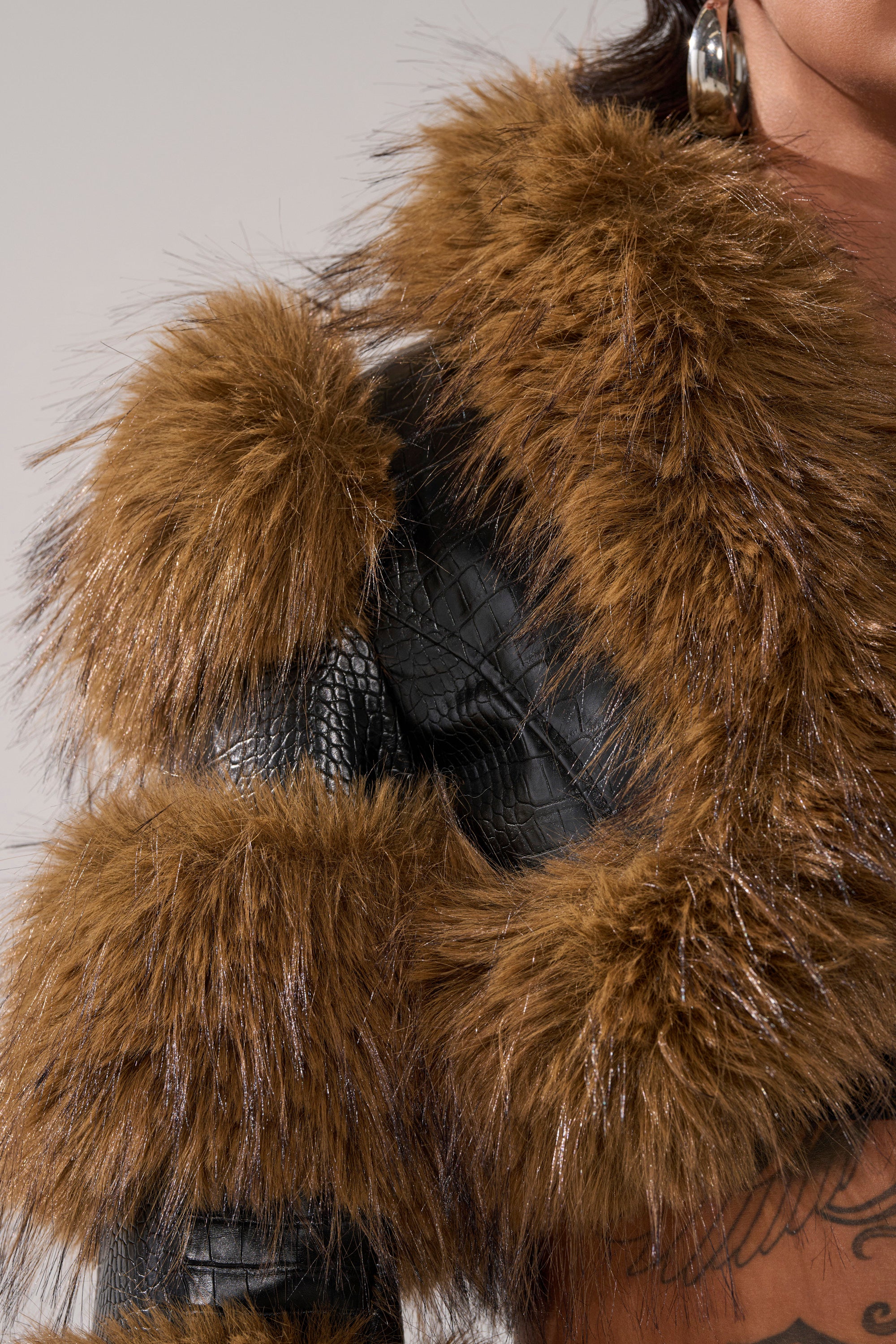 A close-up of someone in the NEW LOVERGIRL ULTRA CROP FUR TRIM MOTO, a black textured crop jacket with thick brown faux fur trim. They accessorize with large hoop earrings and have chest tattoos visible.