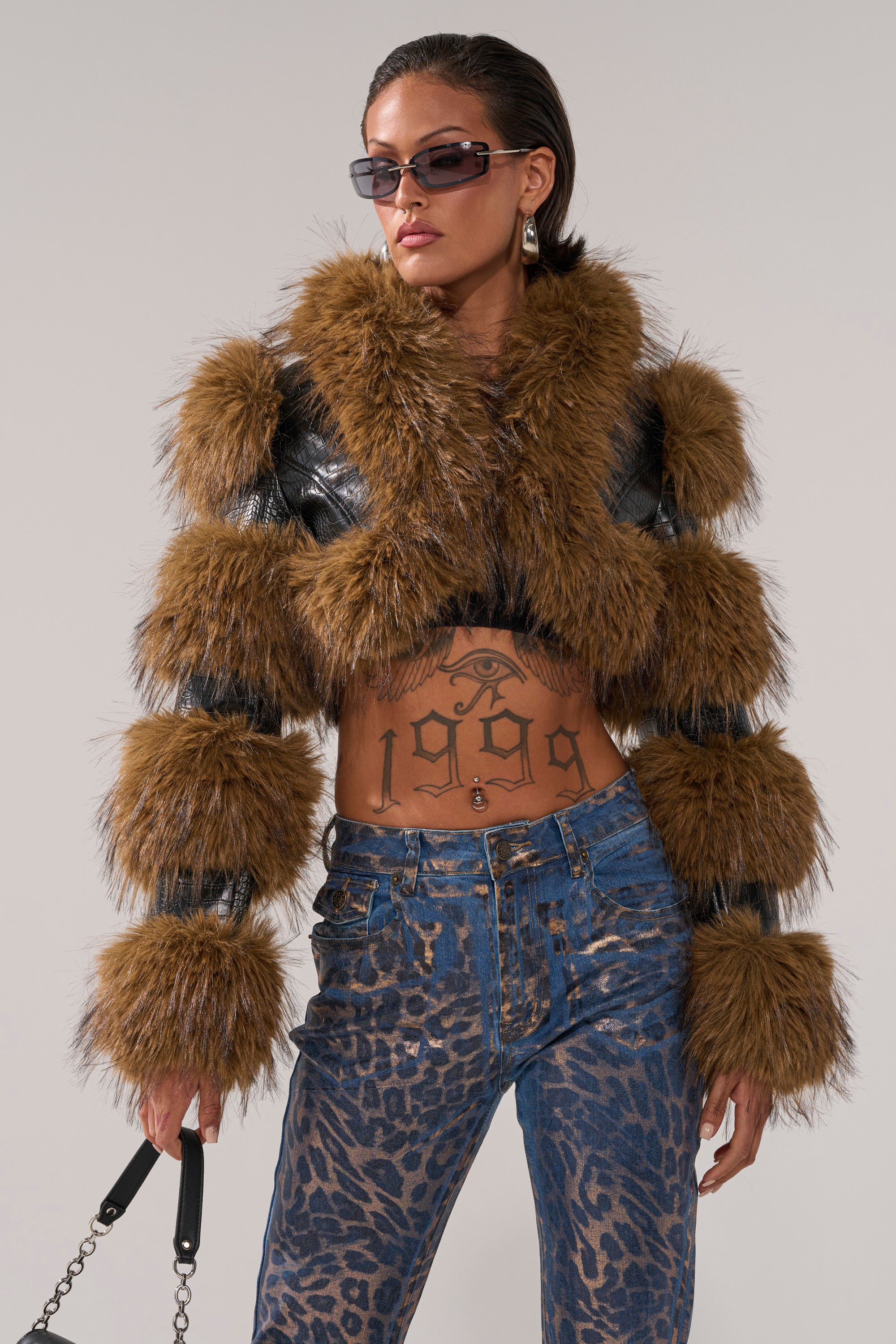 A woman wearing sunglasses, hoop earrings, blue leopard-print pants, and the NEW LOVERGIRL ULTRA CROP FUR TRIM MOTO jacket. She has a visible “1969” stomach tattoo and is holding a strap in one hand.