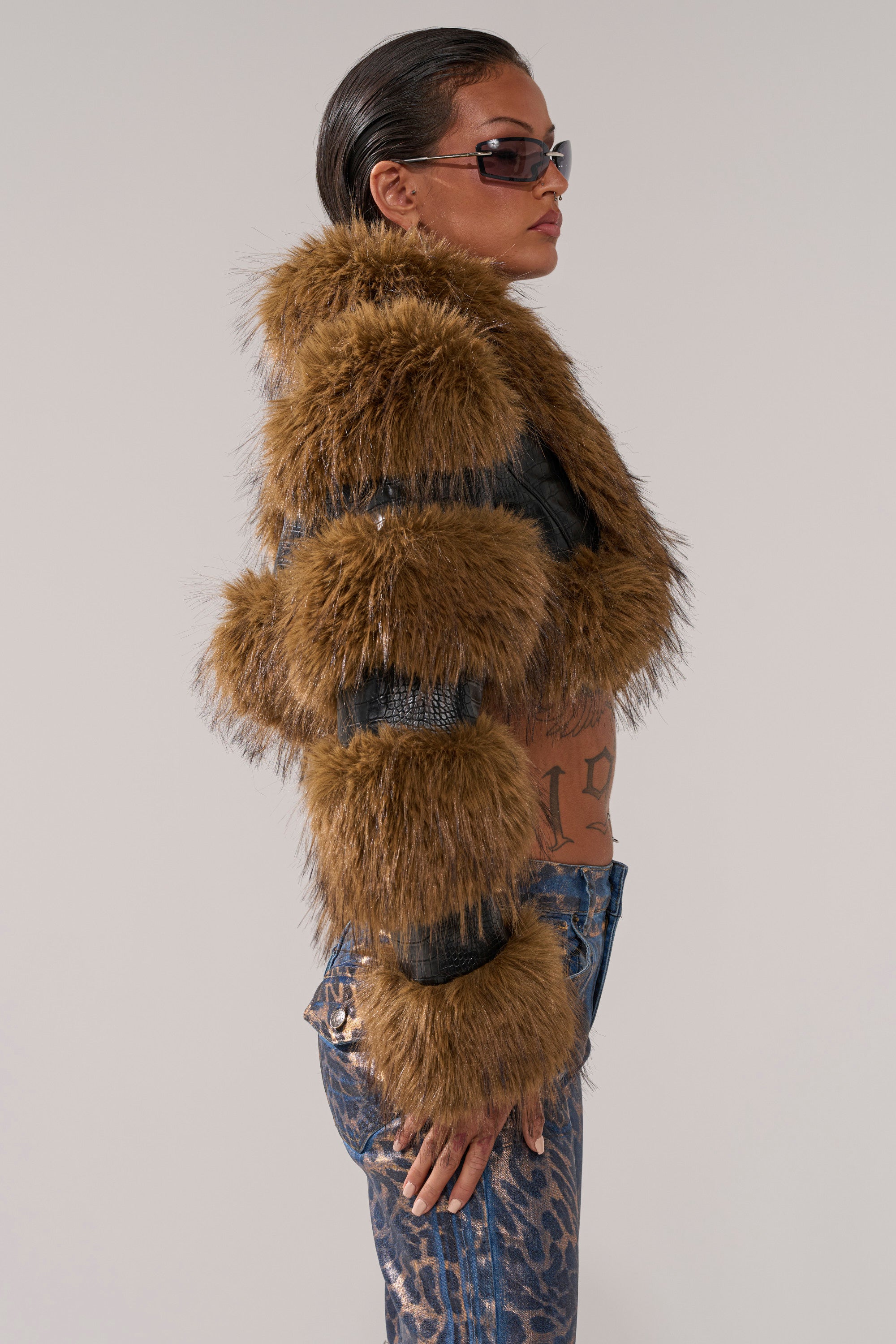 A tattooed individual with slicked-back hair and sunglasses models the NEW LOVERGIRL ULTRA CROP FUR TRIM MOTO jacket and patterned jeans in profile against a plain backdrop—delivering an edgy winter fashion statement.