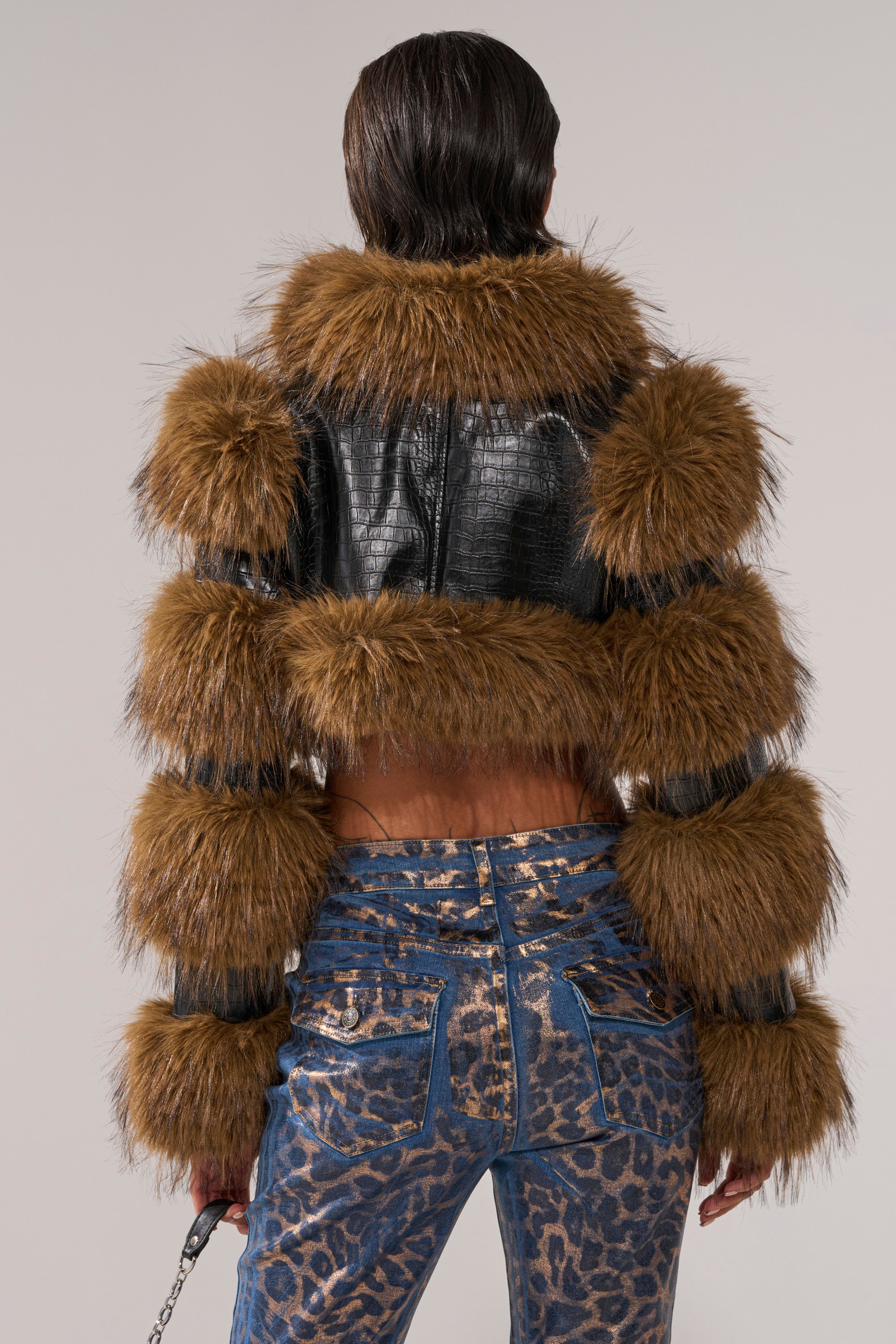 A person faces away, wearing the NEW LOVERGIRL ULTRA CROP FUR TRIM MOTO jacket with fur bands on arms and collar, paired with textured blue embossed snakeskin print jeans, and holding a handbag by their side.