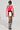 A woman poses confidently against a simple backdrop, modeling the REV IT UP RACING MOTO IN RED jacket. The jacket features embroidered patches and comes with a coordinating mini skirt. Her look is finished with black and white lace-up boots and sunglasses, giving off a daring and fashionable vibe.