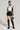 A person stands against a plain background wearing the white "REV IT UP RACING MOTO JACKET" with patches, a black faux leather mini-skirt, and black knee-high lace-up boots. They hold a small green handbag and have their hair slicked back in a polished, sleek style.