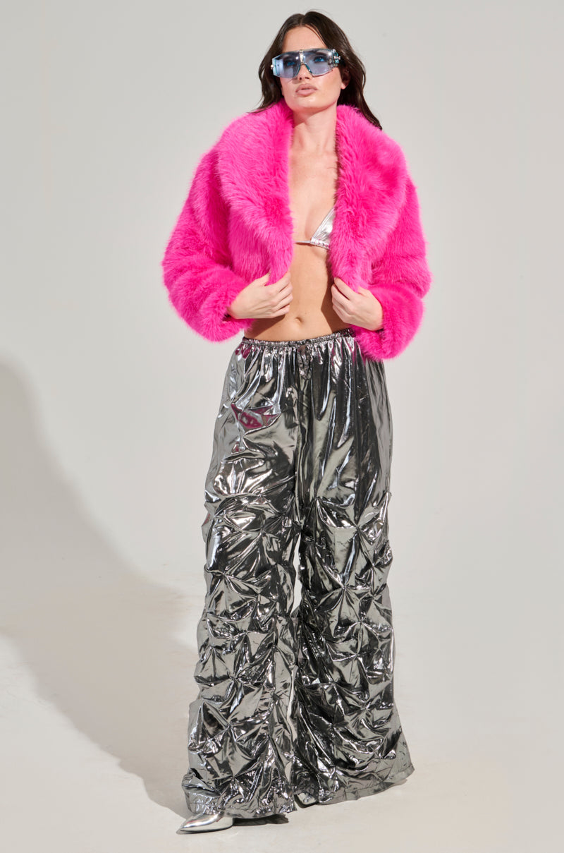 A person confidently stands in the "Lights Out Cropped Faux Fur Jacket in Pink," featuring an oversized collar, alongside a silver bikini top and shiny silver pants. Their long dark hair flows as reflective sunglasses complete the bold look against a plain background.