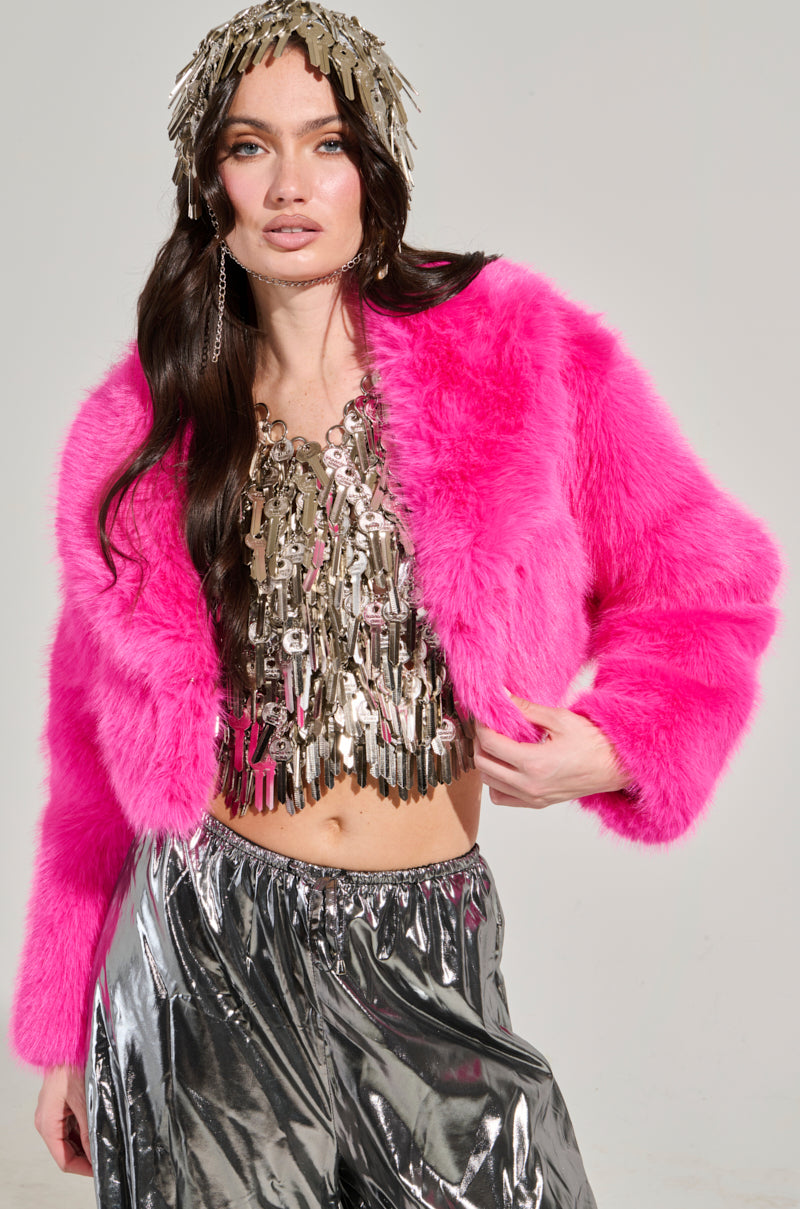 A woman poses against a plain background in the LIGHTS OUT CROPPED FAUX FUR JACKET IN PINK, paired with a metallic fringe top and shiny silver pants. Her long dark hair complements the headpiece made of metallic tassels, adding an eclectic charm to her striking outerwear ensemble.