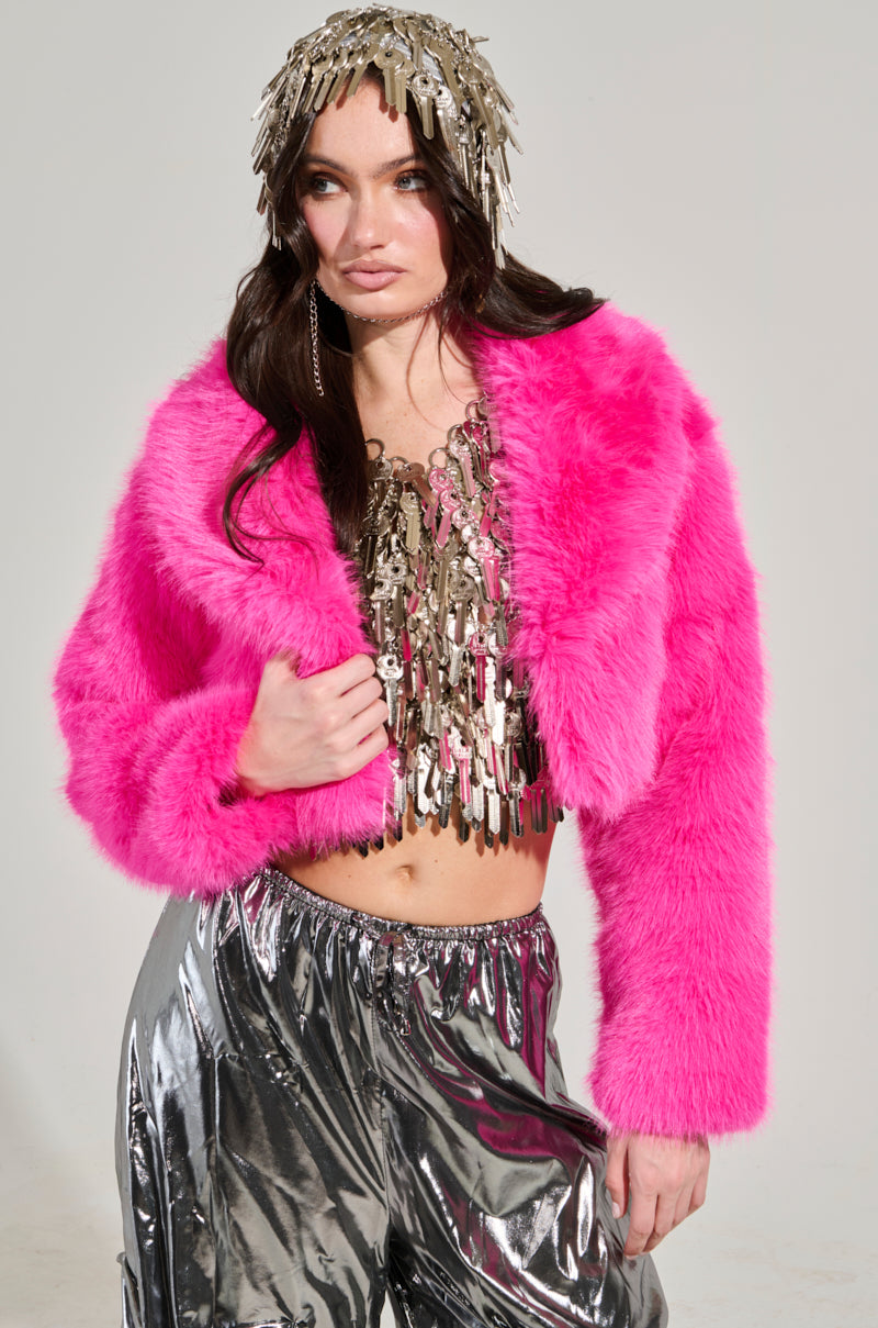A person poses in the LIGHTS OUT CROPPED FAUX FUR JACKET IN PINK, layered over a shimmering metallic top with fringe details. They wear glossy silver pants and a headpiece adorned with silver tassels, standing against a plain background, showcasing a chic cropped silhouette.