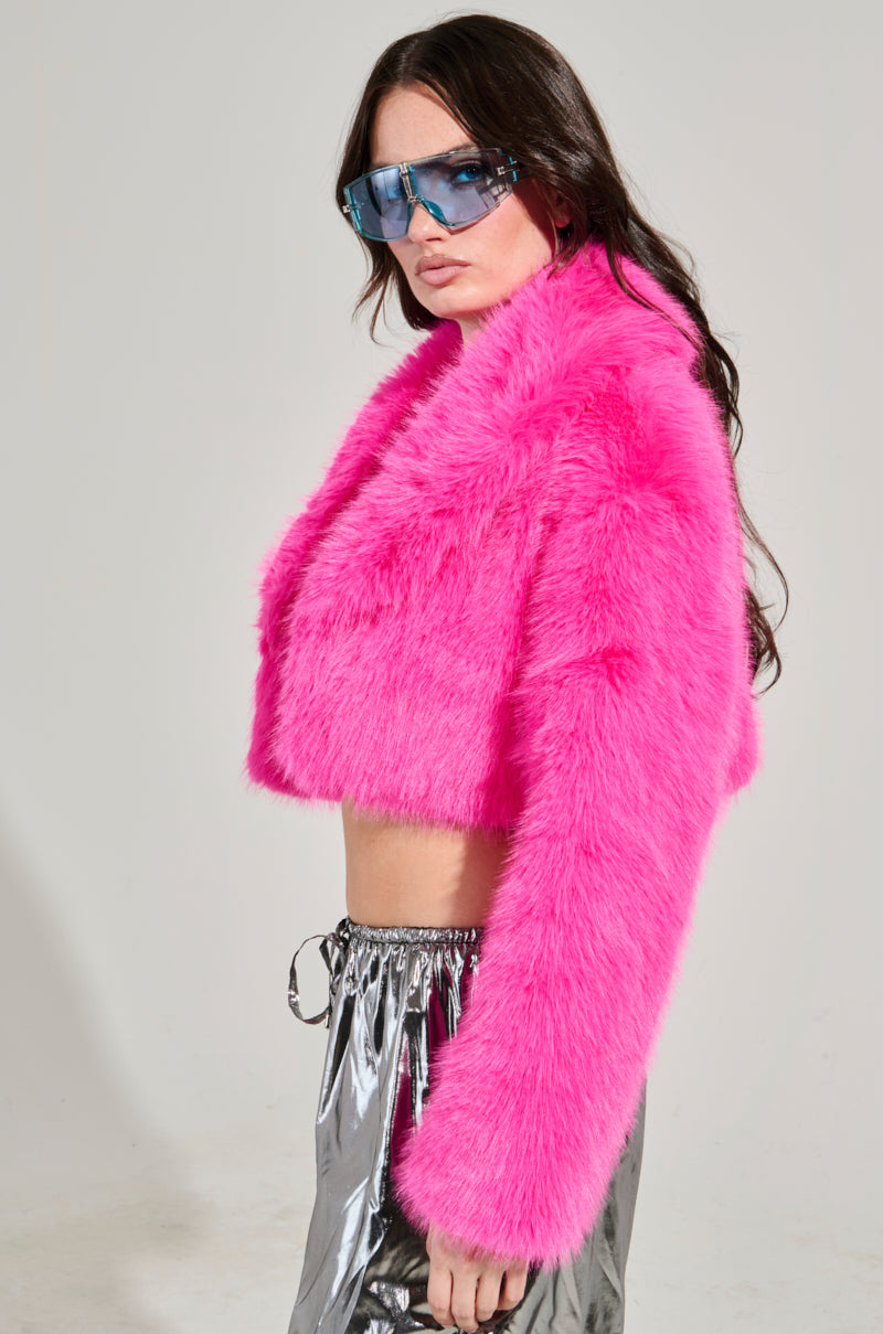 A person confidently poses in front of a plain background, wearing the LIGHTS OUT CROPPED FAUX FUR JACKET IN PINK with metallic silver pants and blue-tinted sunglasses. Their long, dark hair cascades over the vibrant jacket.