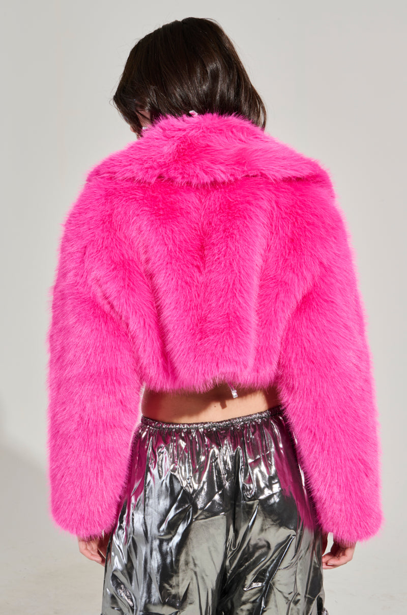 The individual is facing away from the camera, highlighting the LIGHTS OUT Cropped Faux Fur Jacket in Pink, paired with shiny silver metallic pants. The plain, light-colored background accentuates the vibrant outfit.