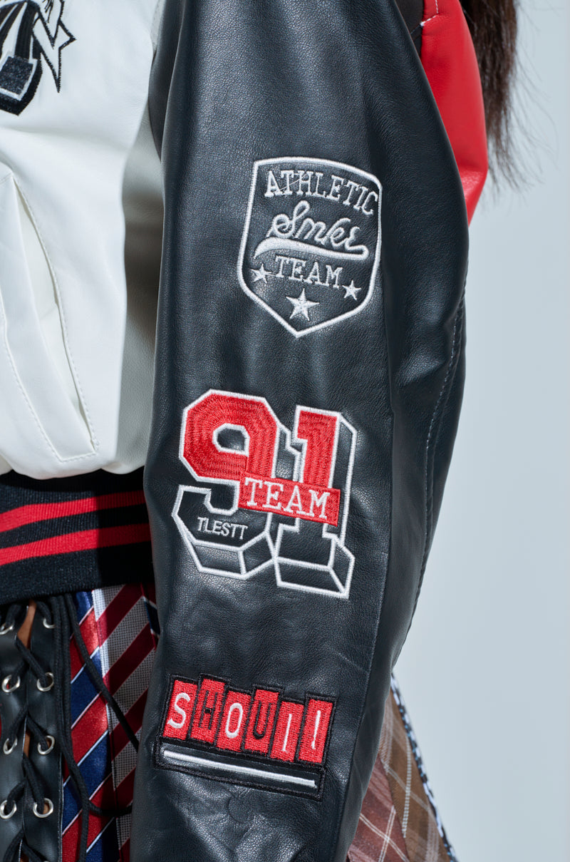 Close-up of the Champions League Faux Leather Bomber sleeve, adorned with sports-themed patches, including a shield reading "ATHLETIC EST 1959 TEAM," numbers "91 TEAM," and a red patch with "SHOUT." The bomber style pairs well with the partially visible striped and plaid skirt.