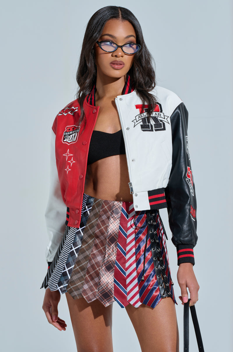 A woman stylishly poses against a plain backdrop, wearing the CHAMPIONS LEAGUE FAUX LEATHER BOMBER with sports-themed patches, a black crop top, and a geometric patterned skirt. She has cat-eye glasses, loose wavy hair, and holds a bag in her right hand.