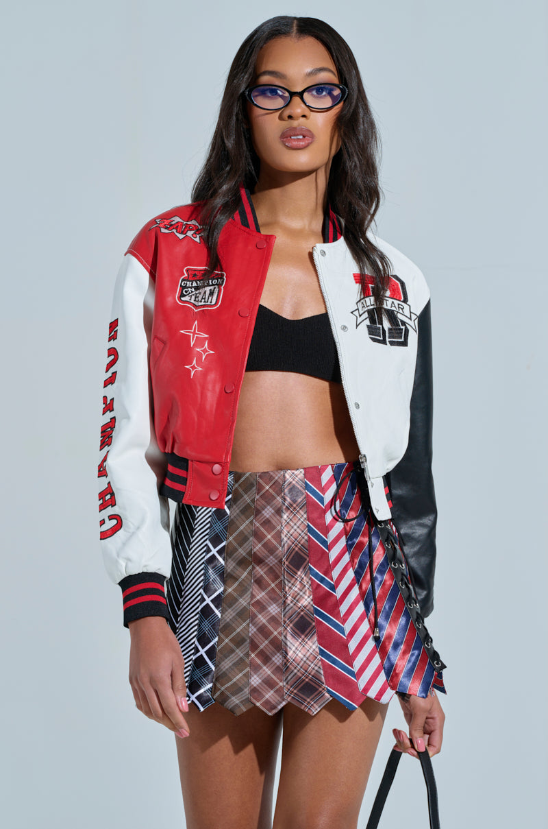 A woman in a CHAMPIONS LEAGUE FAUX LEATHER BOMBER, a black bandeau top, and a skirt of colorful neckties, with long dark hair and glasses, stands against a light blue background.