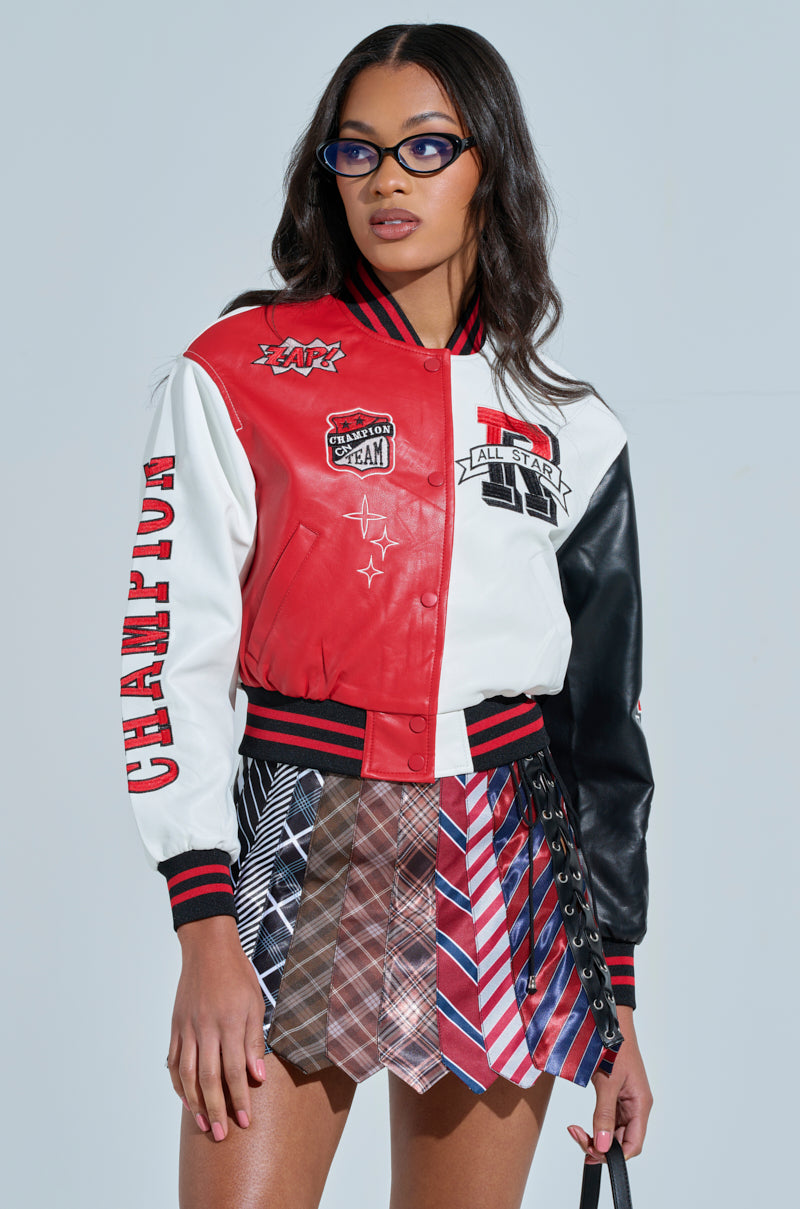 A person wears the CHAMPIONS LEAGUE FAUX LEATHER BOMBER, a red and white varsity jacket with sports-themed patches, along with a skirt made from colorful ties. Black glasses add to the look, which boasts star elements and bold text details.