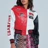 A person wears the CHAMPIONS LEAGUE FAUX LEATHER BOMBER, a red and white varsity jacket with sports-themed patches, along with a skirt made from colorful ties. Black glasses add to the look, which boasts star elements and bold text details.