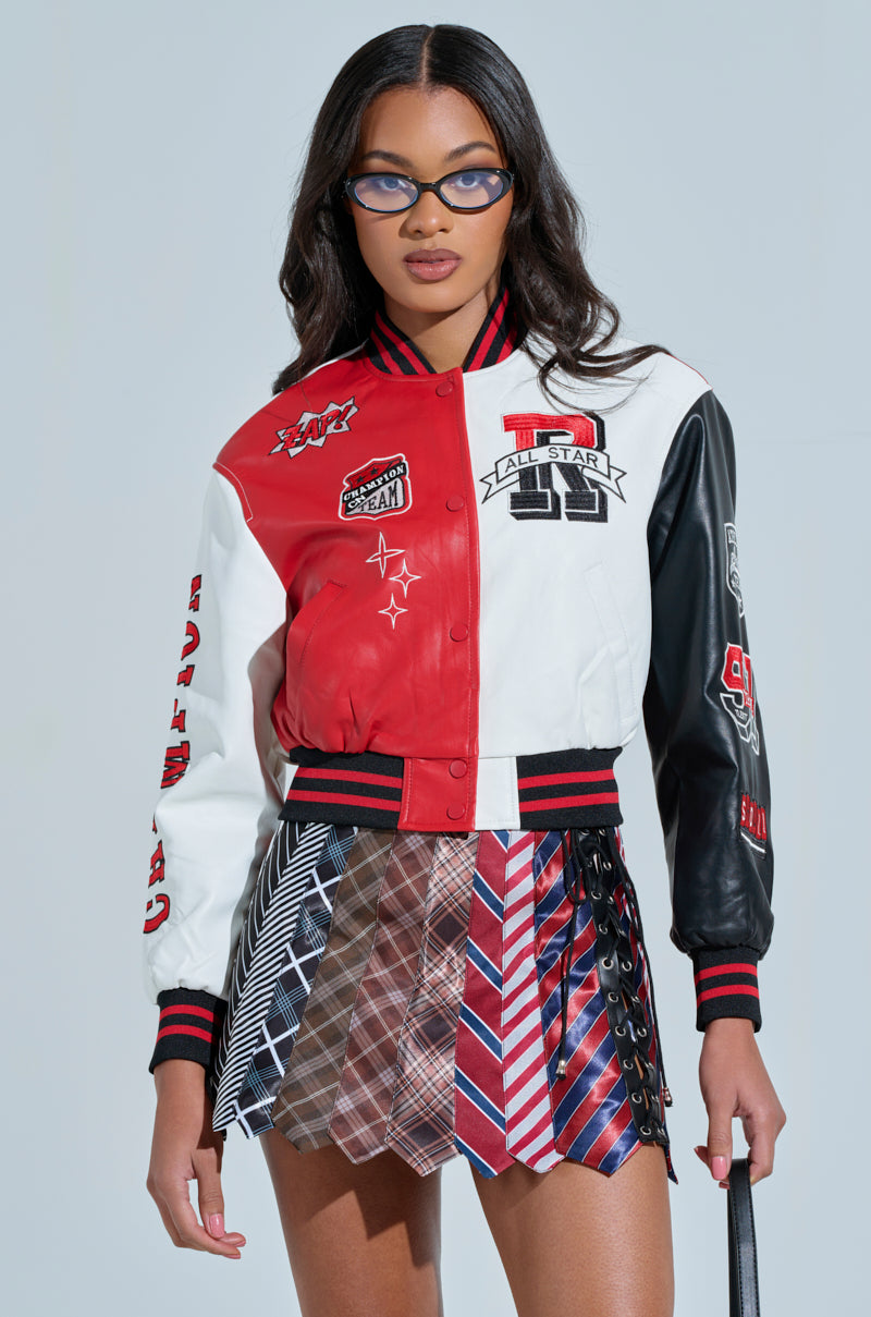 A person wears the CHAMPIONS LEAGUE FAUX LEATHER BOMBER in a red, white, and black color block design with sporty patches, paired with a unique skirt of patterned neckties and glasses. The backdrop is plain light blue.