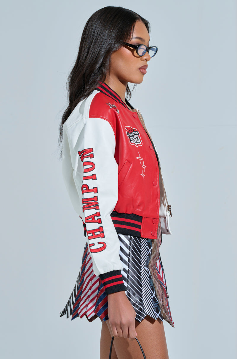 Woman in glasses wearing the CHAMPIONS LEAGUE FAUX LEATHER BOMBER, red and white with "Champion" on the sleeve, paired with a colorful plaid skirt. She holds a sports patch-adorned black bag against a light backdrop, facing left.