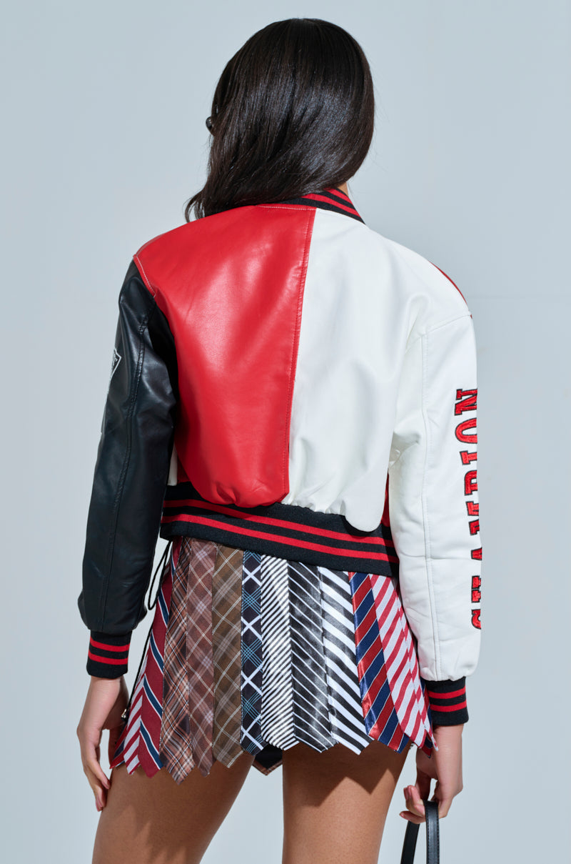 A person with long hair sports a red, white, and black CHAMPIONS LEAGUE FAUX LEATHER BOMBER adorned with sports-themed patches and a text logo on the sleeve, paired with a pleated plaid skirt as they face away from the camera.