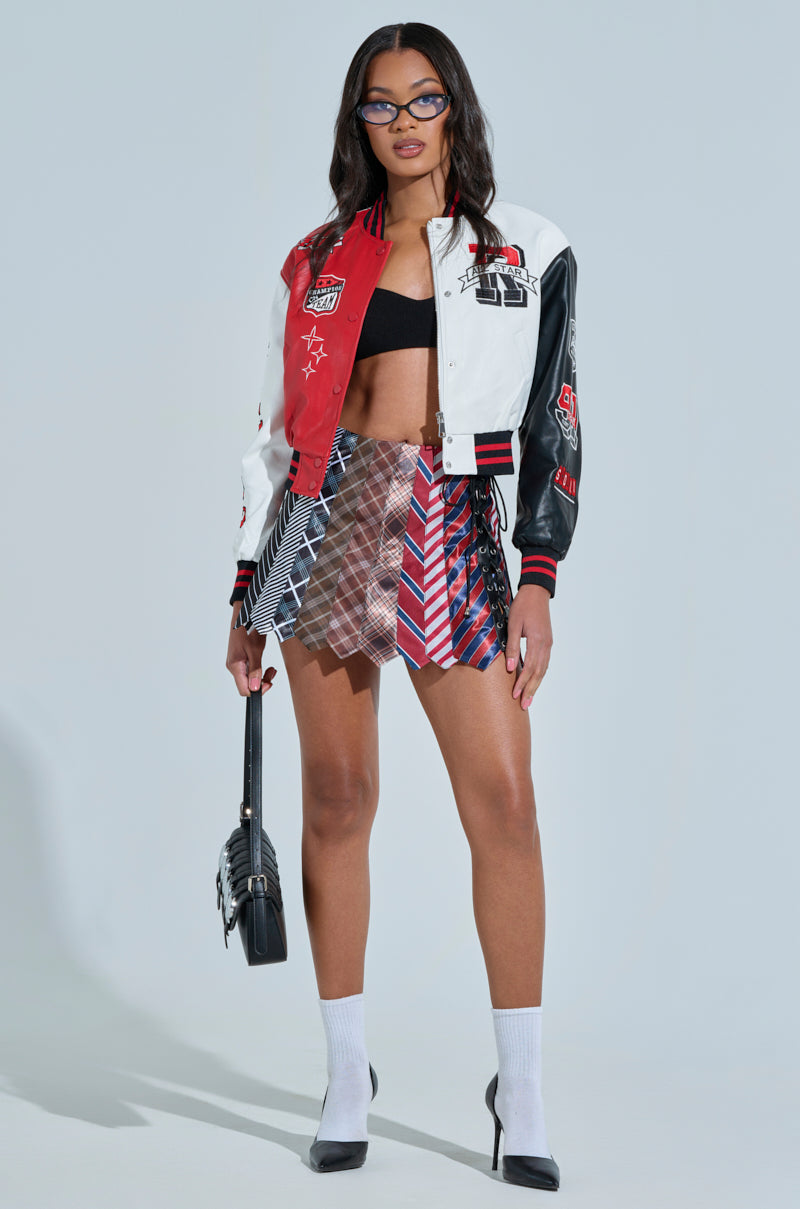 A person wearing glasses, a CHAMPIONS LEAGUE FAUX LEATHER BOMBER with sports-themed patches, a black crop top, and a patterned mini skirt. They're holding a small black handbag and wearing white socks with black heels, standing in a neutral pose against a plain background.