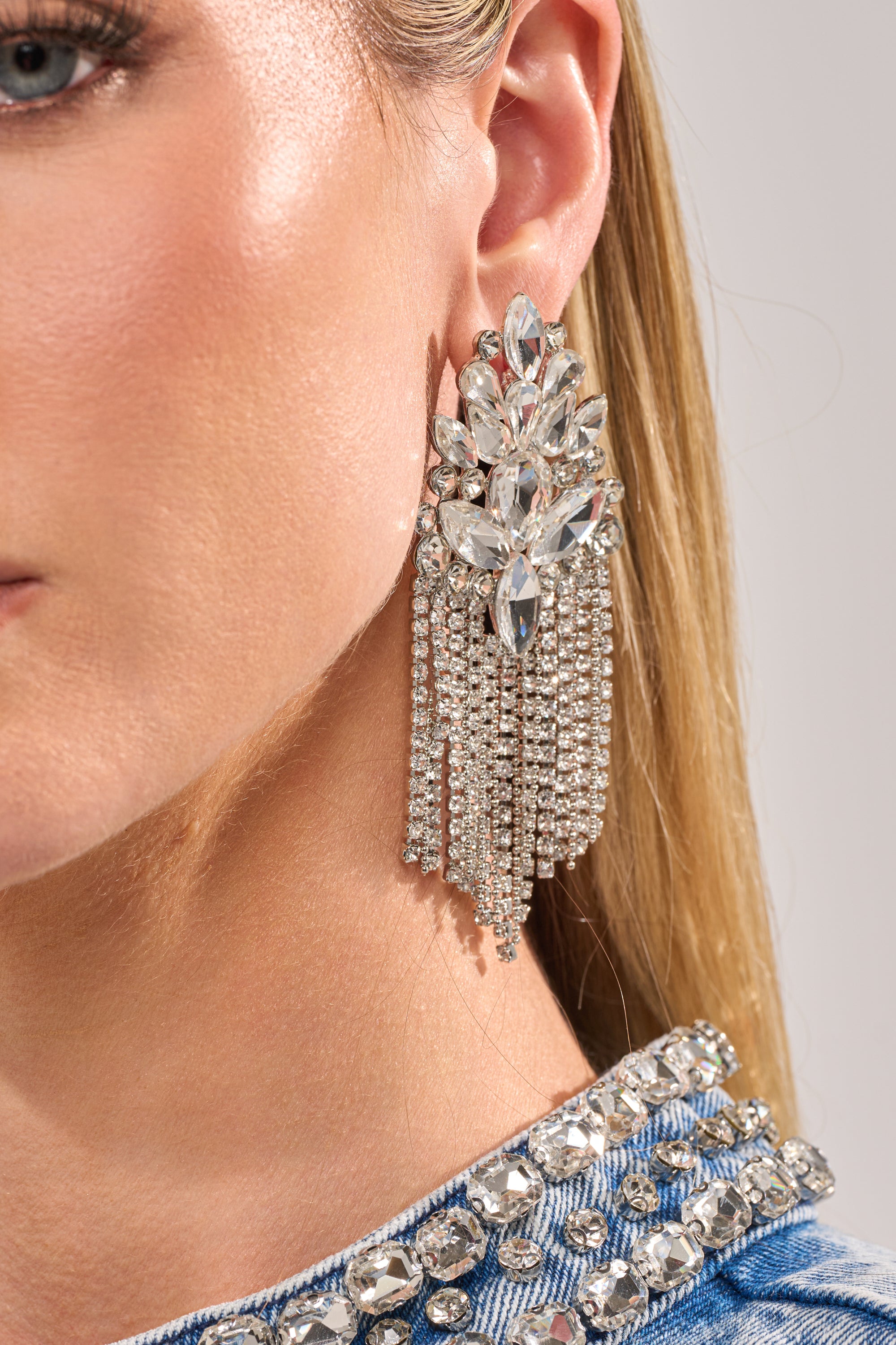 Close-up of a woman's ear wearing GNO EARRING—large rhinestone cluster earrings with clear stones and fringe chains. She has straight blonde hair and a studded denim top, making these earrings perfect for wedding guest accessories. 1