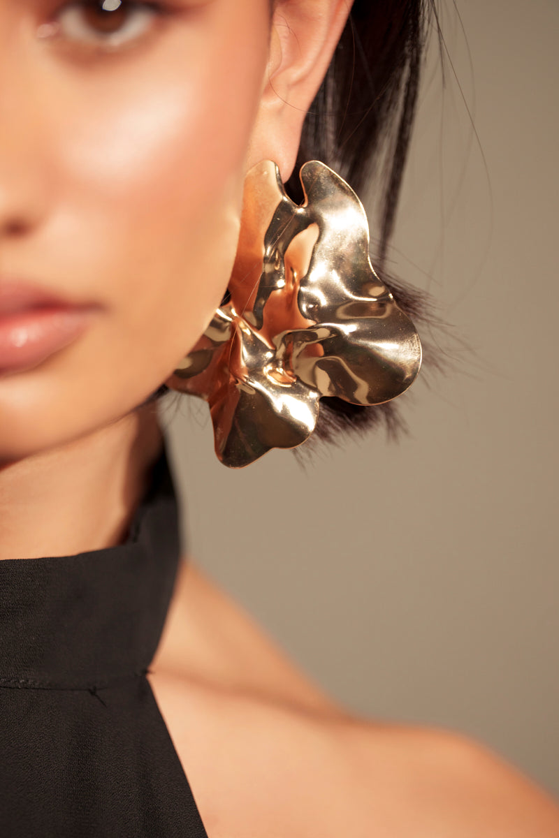 Close-up of a woman in a black halter top with dark hair tucked behind her ear, wearing the SPREAD YOUR WINGS EARRING—its metallic faux gold, abstract wavy flower silhouette stands out beside her face.