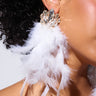 A close-up image captures a person with curly hair and a small ear piercing, adorned with the EMPOWER FEATHER EARRING—a luxurious statement accessory featuring sparkling stones and elegant white feathers. The plain, neutral background enhances the lobe posts embellished with gemstones.