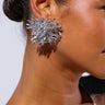 A person with dark, smooth skin is shown from the side wearing an elaborate, spiky METAL MANIA EARRING crafted in faux silver metal. Their hair is styled in a sleek bun. The top they've donned is partially visible, showcasing a mix of lace and a pink and black pattern. The background is plain white.