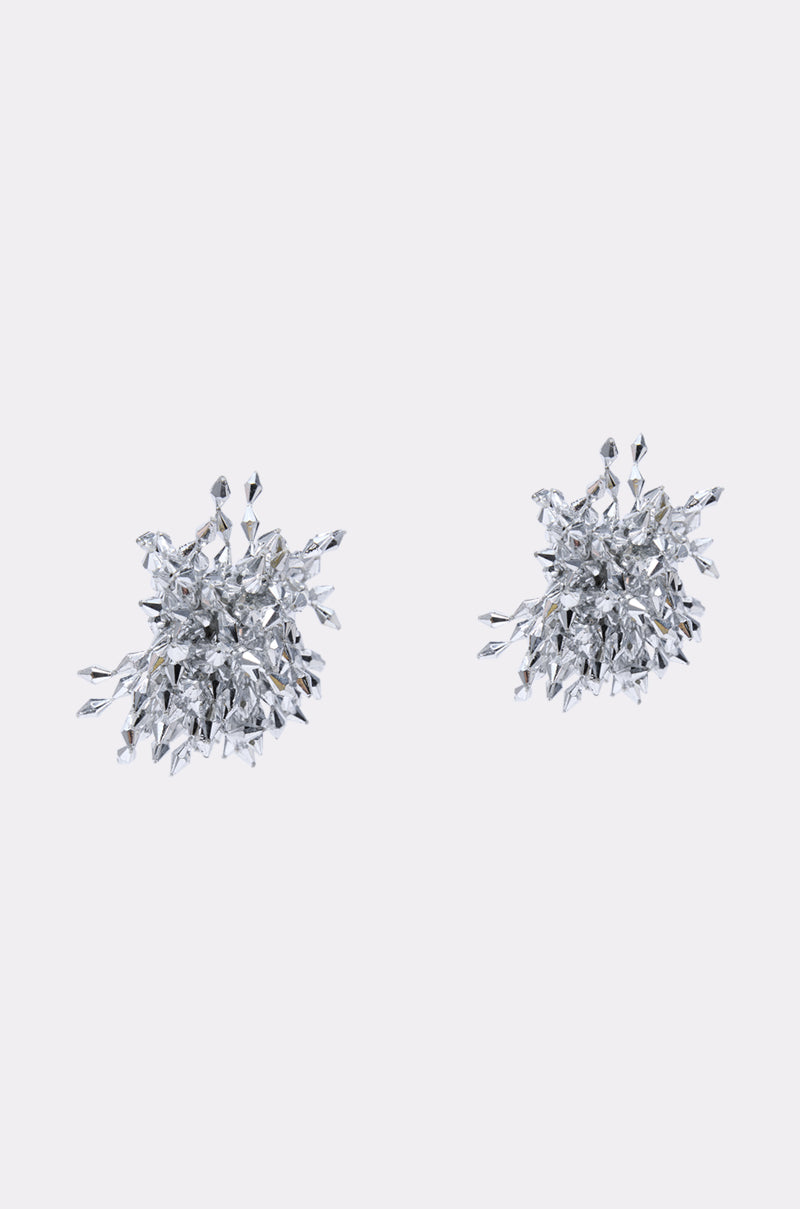The METAL MANIA EARRING features intricate silver starburst shapes with diamond embellishments, their sparkling, pointed crystal-like details standing out beautifully against a plain light grey background.