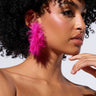 A woman with curly hair wears striking pink Feather Fury Hoop earrings. She is turned slightly to the side, her hand gently touching her cheek. Dressed in a strapless black top against a plain, light background, she exudes an elegant and fashionable aura accentuated by metallic faux gold accents.