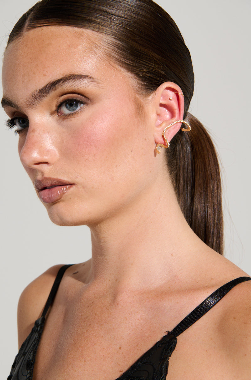A woman with long, dark hair in a sleek ponytail is wearing a black strappy top. Her ear is adorned with a metallic faux gold snake-shaped cuff and the COUTURE WRAP EARRING, which adds an elegant touch. The light-colored, plain background accentuates her chic accessories. 0