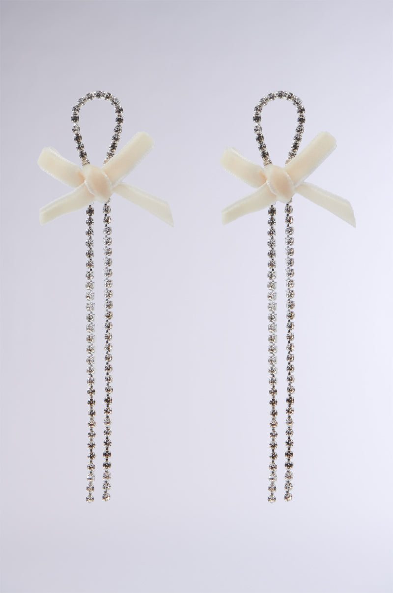 The BOW BABE EARRING features two elegant pieces with velvet bows at the top, showcasing delicate dangling chains embellished with sparkling crystals. The design is both sophisticated and graceful, set against a plain light gray background.