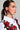 A model with straight brown hair in a sleek ponytail is shown in profile. She wears a white blouse adorned with red embroidered roses and green leaves. A red beaded ribbon decorates her left ear, resembling the TREND DRIVEN BOW EARRING by AKIRA Label. The background is plain gray.