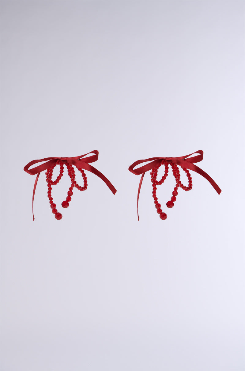 The image showcases TREND DRIVEN BOW EARRINGS, featuring two striking red pieces with an oversized bow design, complete with ribbon strands and dangling bead strings. These statement earrings are elegantly positioned side by side against a plain light background.