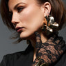 A woman with short brown hair poses in a side profile, highlighting her standout ORGANIC VIBES EARRING. She is adorned in a black lace glove and a black outfit. Her serious expression against the muted gray background enhances the earring's metallic faux gold charm.