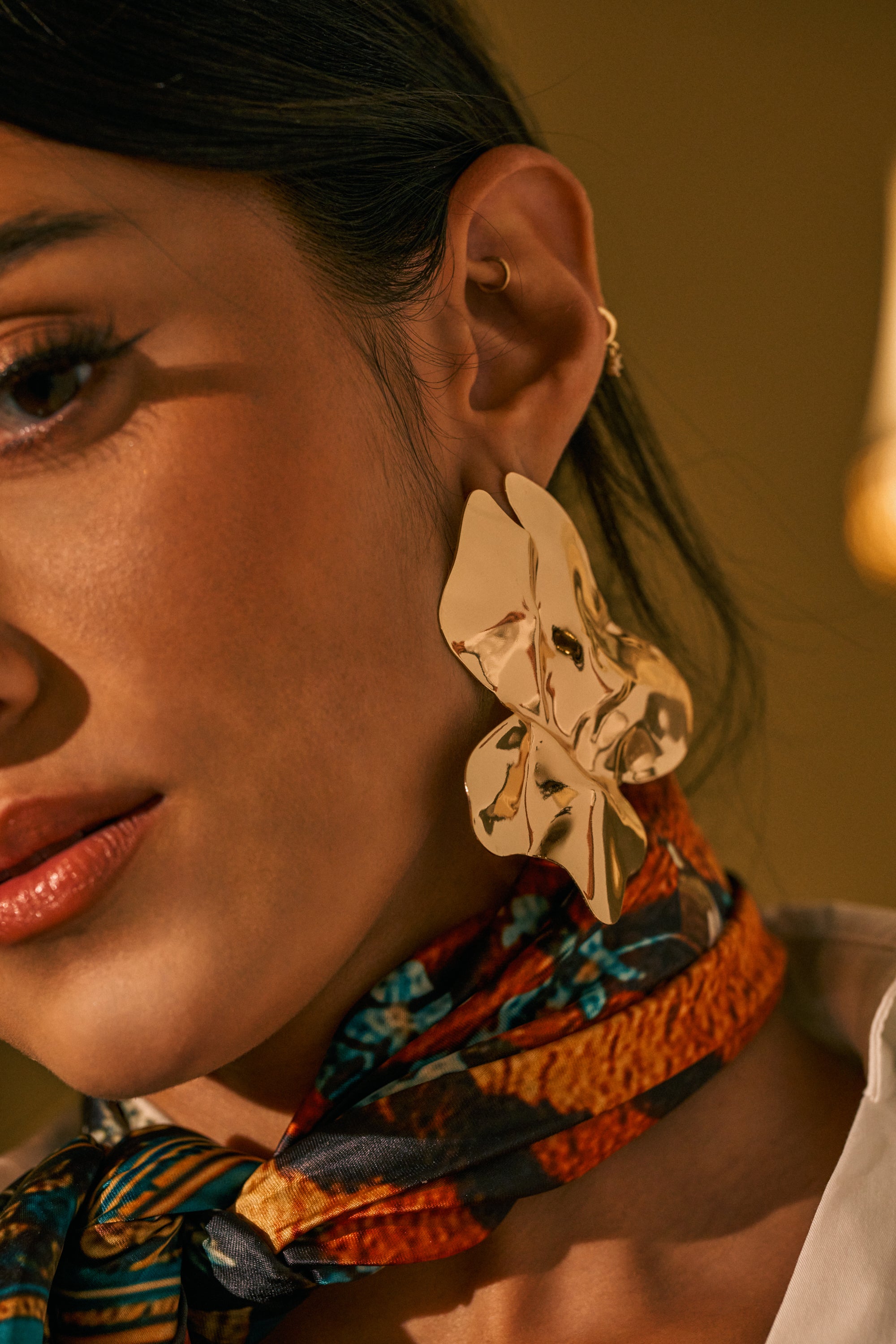 Close-up of a woman wearing the ORGANIC VIBES EARRING—a large, sculptural gold statement piece—paired with a colorful scarf around her neck, bathed in warm lighting with a gentle, soft focus on her face.