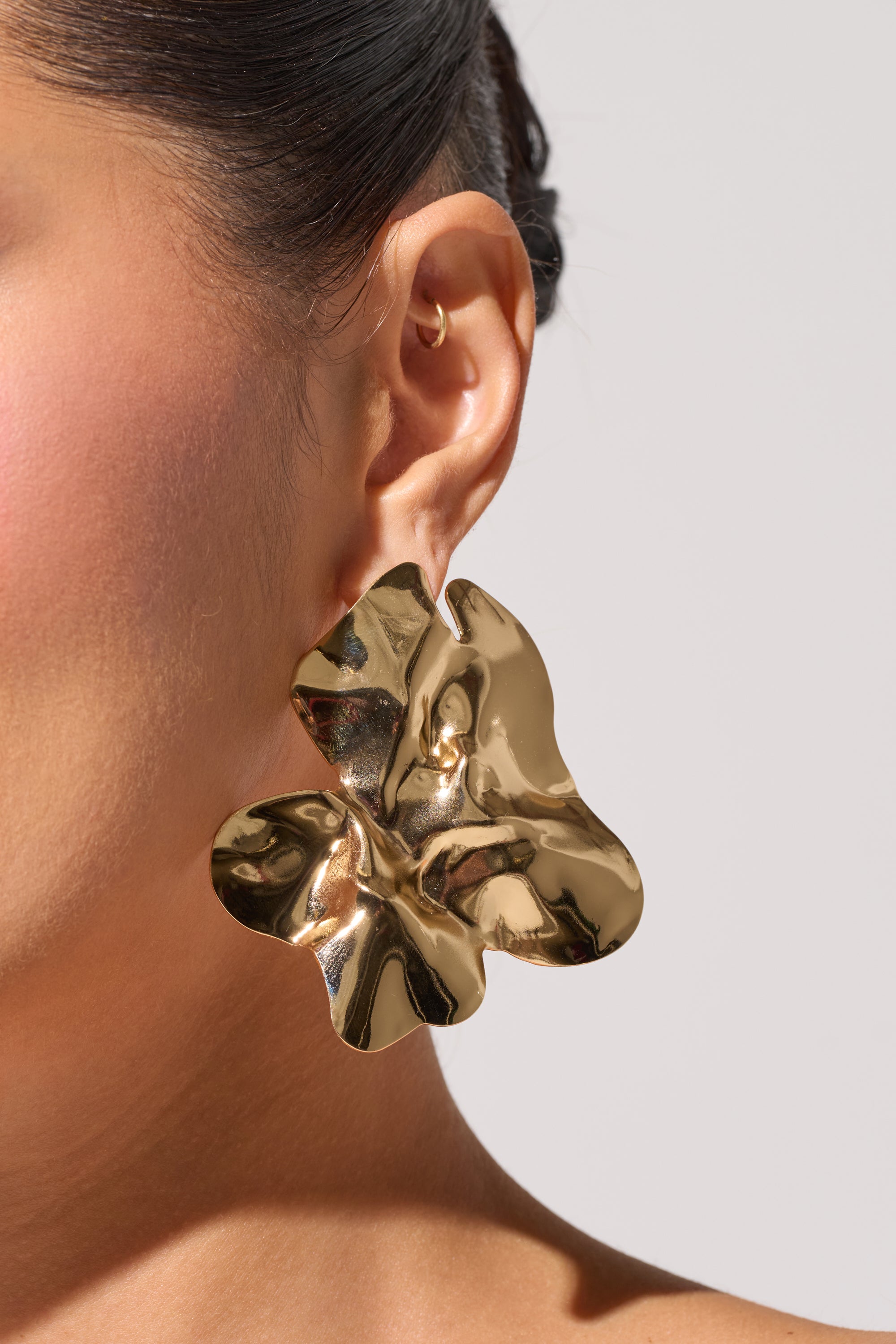 Close-up of a person's ear adorned with the ORGANIC VIBES EARRING, a large, shiny gold statement piece featuring an abstract design. The background is neutral, with dark hair pulled back and part of the face visible. 1