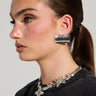 A woman with sleek, dark hair tied back is wearing large, rectangular MOD EARRING in metallic faux silver and a sparkling floral necklace. She is dressed in a black top with small white text. The background remains neutral, enhancing her minimalist design.