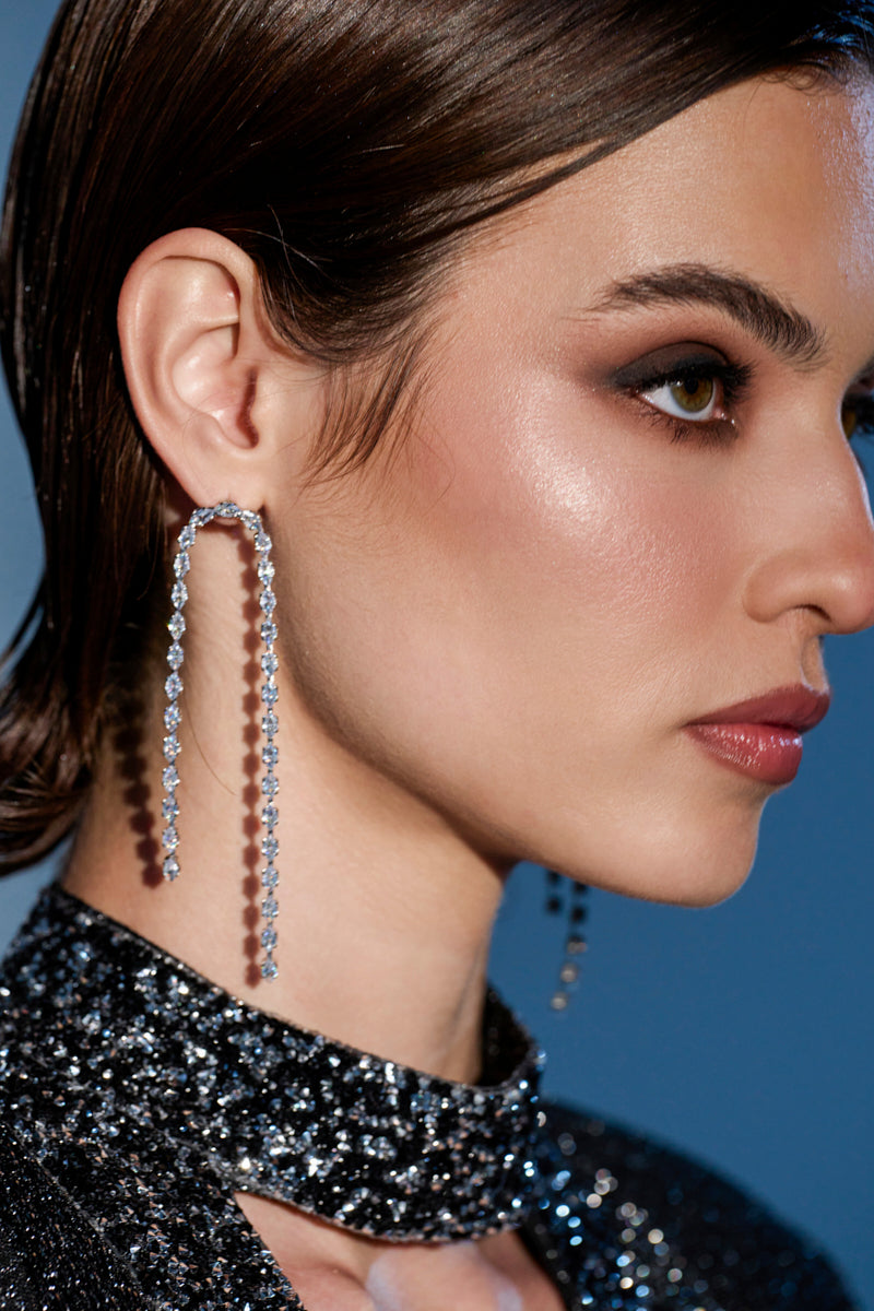 A close-up of a woman with sleek brown hair, dramatic makeup, and a sparkly high-neck top showcases the WATERFALL DANGLE EARRING IN SILVER, featuring glittering gems, set against a blue background. 0
