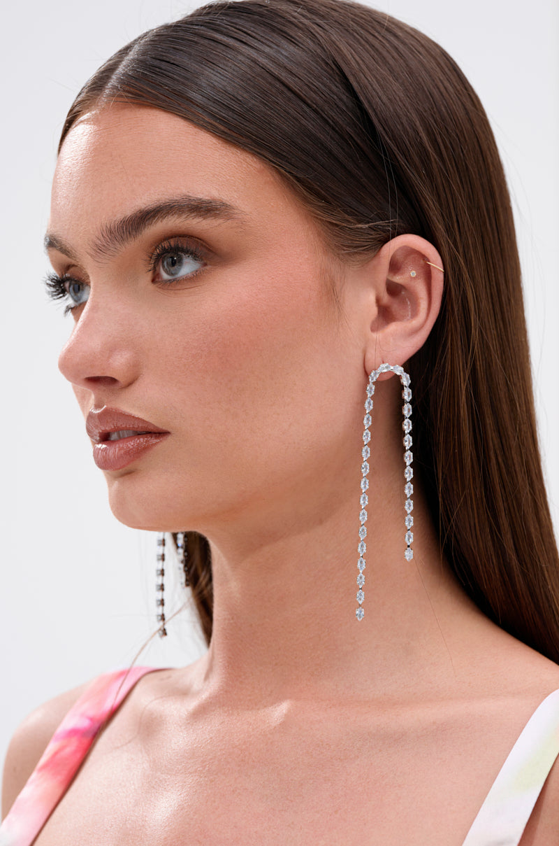 A woman with straight brown hair and natural makeup wears the WATERFALL DANGLE EARRING IN SILVER, featuring long, metallic dangles accented with clear gemstones. She pairs them with a colorful sleeveless top against a plain background. 2
