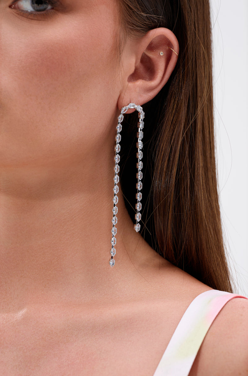 A woman with straight brown hair wears the WATERFALL DANGLE EARRING IN SILVER, a long, silver accessory with clear gemstones that curves from her earlobe and drops below her shoulder. Only her profile and one shoulder are visible. 4