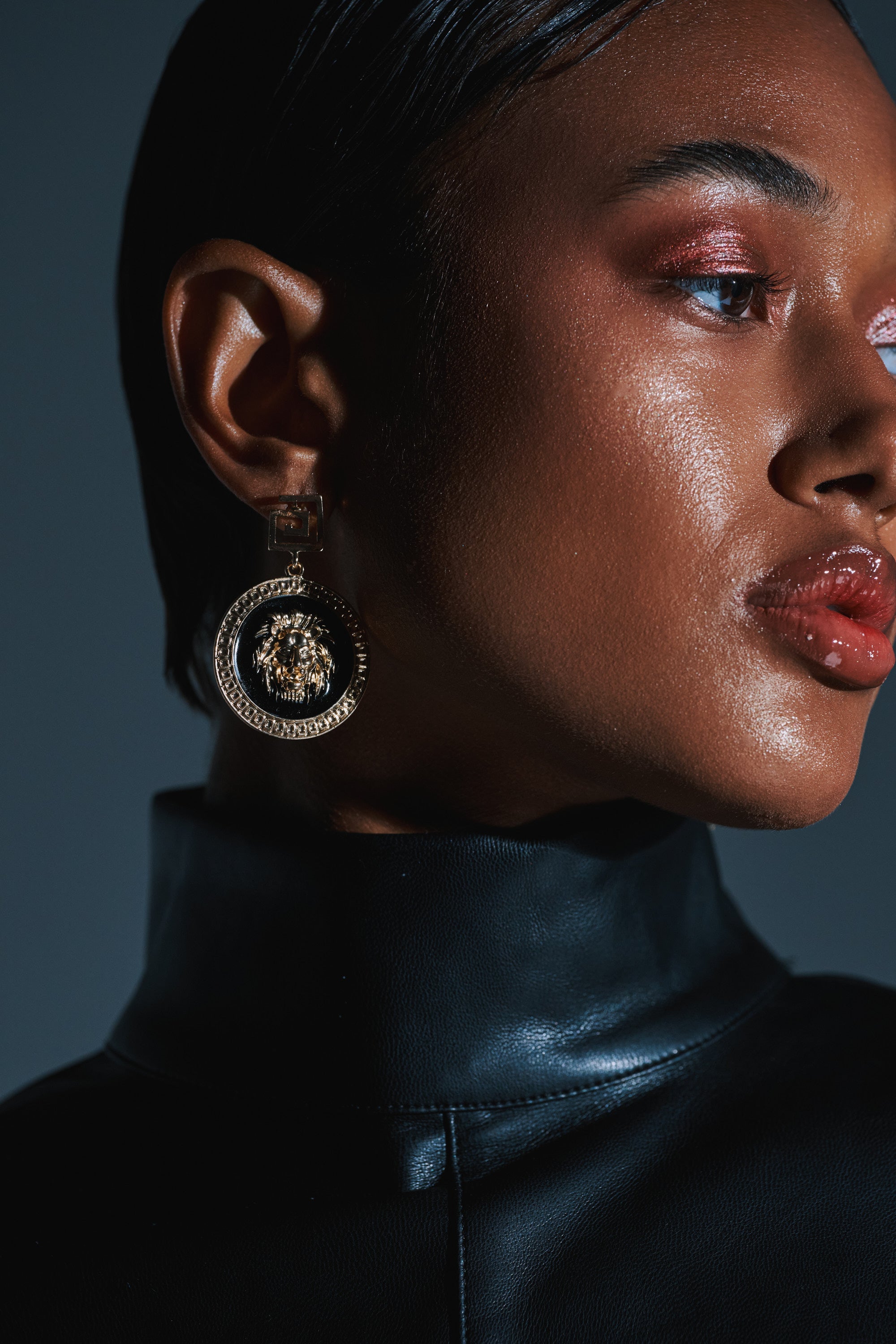 A close-up of a person with glossy skin and slicked-back hair, wearing a shiny black leather turtleneck and the bold LION'S SHARE EARRING, plus shimmery eyeshadow and glossy lips. 1