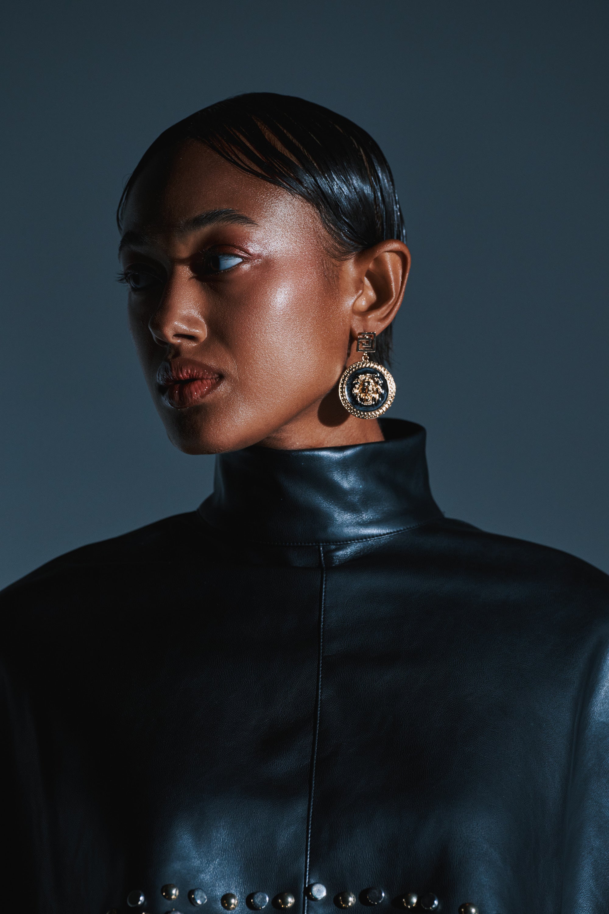 A woman with sleek, pulled-back hair gazes to the side against a dark background, highlighting her bold LION'S SHARE EARRING and a high-neck black leather top adorned with metallic studs. 2