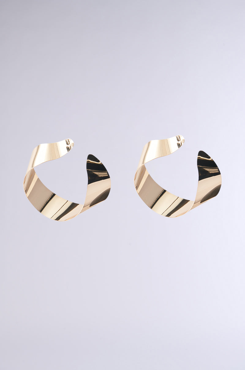 The BENT OUTTA SHAPE EARRING features twisted faux gold metallic hoops with a sleek, modern design from the AKIRA Label. The shiny finish beautifully captures light, adding a stylish and sophisticated touch against a light gray background. 0