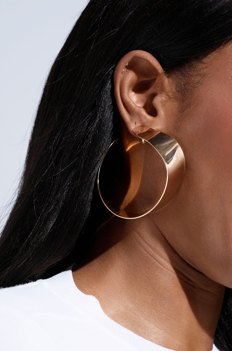 Close-up of someone with long dark hair wearing the AKIRA Label's BENT OUTTA SHAPE EARRING. The large, chunky open hoop in faux gold metal reflects light brilliantly. They are in a white shirt, and the image focuses on their ear and the dazzling earring. 1