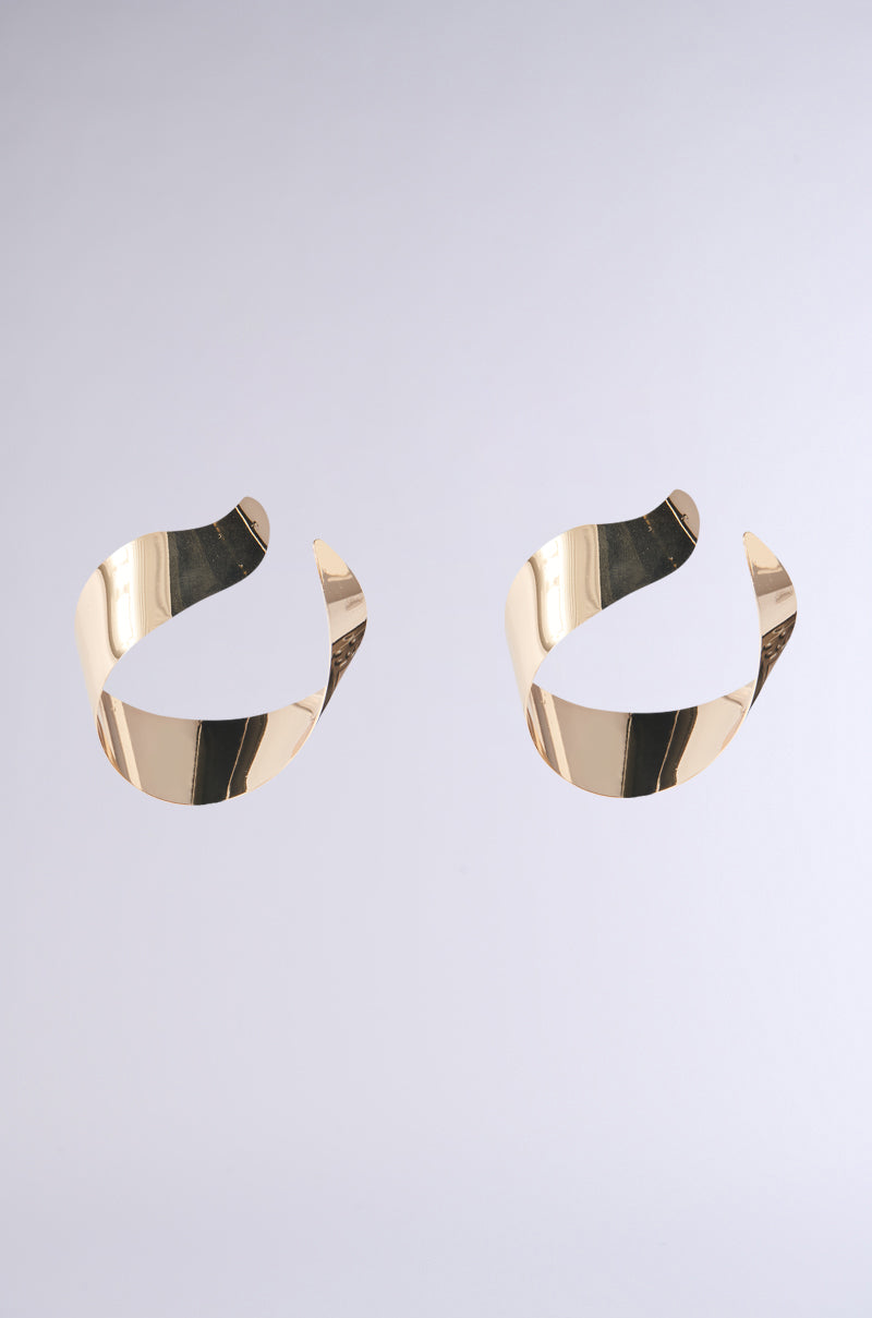 The BENT OUTTA SHAPE EARRING from the AKIRA Label, featuring a faux gold metallic spiral design, is displayed against a light gray background. The chunky open hoop has a smooth, reflective surface and is arranged side by side. 2