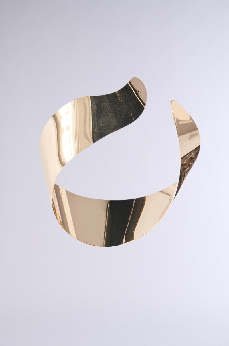 A sleek, shiny faux gold metallic ribbon forms a twisting loop against a gradient gray background, giving the impression it is floating in mid-air, akin to a BENT OUTTA SHAPE EARRING. 3