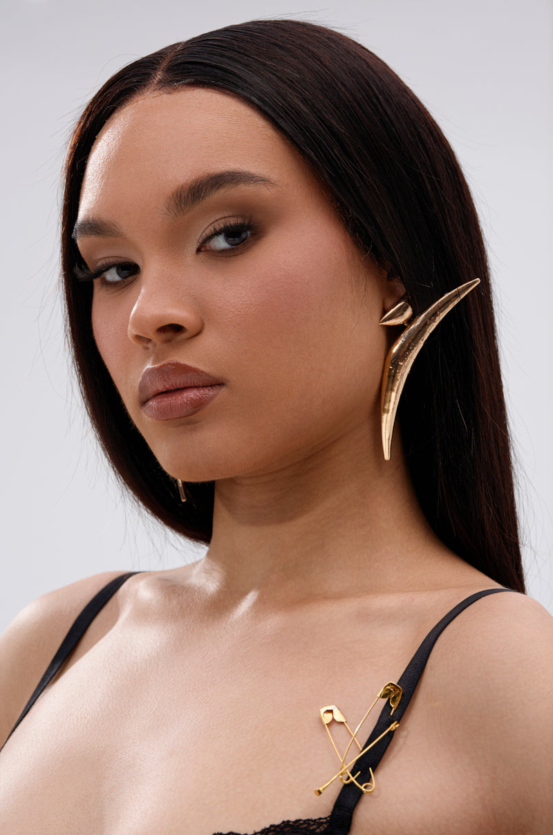 A woman with straight dark hair wears the bold, sculptural DONT PLAY EARRING in gold and a black top with gold safety pins. Her makeup is neutral and she poses confidently against a plain background. 2
