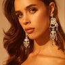A woman with wavy brown hair confidently gazes into the camera, wearing the CHANDELIER DROP EARRING and a strapless red top against a warm, neutral background.