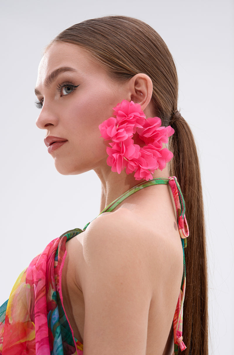 A woman with long brown hair in a sleek ponytail wears the FULL BLOOM HOOP EARRING, featuring floral appliques, paired with a colorful floral-patterned halter top. She gazes to the side against a plain light background. 1