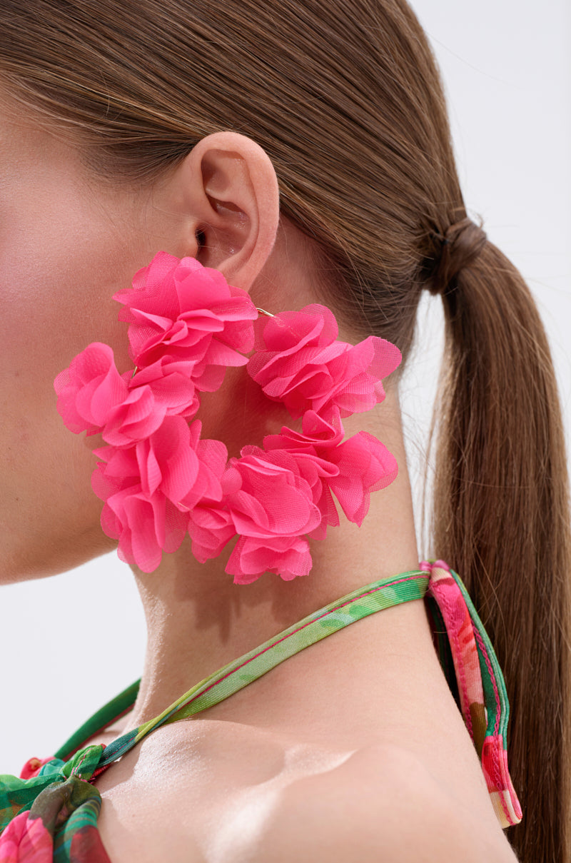 Close-up of a person with long brown hair in a low ponytail, wearing the FULL BLOOM HOOP EARRING made from organza-like fabric and a floral green halter top, against a plain light background. 2