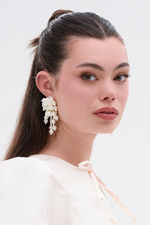 A woman with fair skin and straight brown hair styled half-up wears a white blouse and the REGAL PEARL DROP EARRING, gazing over her shoulder against a light background—an elegant, timeless look ideal for bridal styling. 1