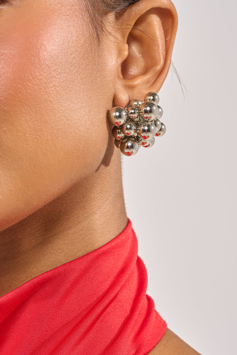 Close-up of a person wearing the BUBBLES AND BRUNCH SILVER EARRING with a red high-neck top, highlighting the contemporary accessory against their cheek and neck. 1