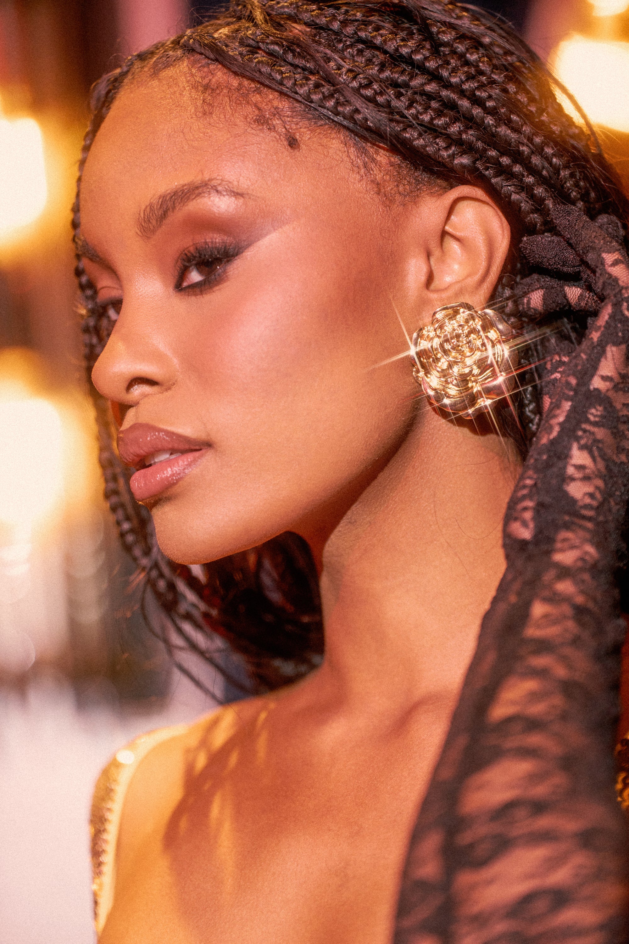 A woman with braided hair and bold makeup gazes to the side, wearing IN BLOOM EARRING. Warm, golden lighting in the background creates a glamorous atmosphere. 1